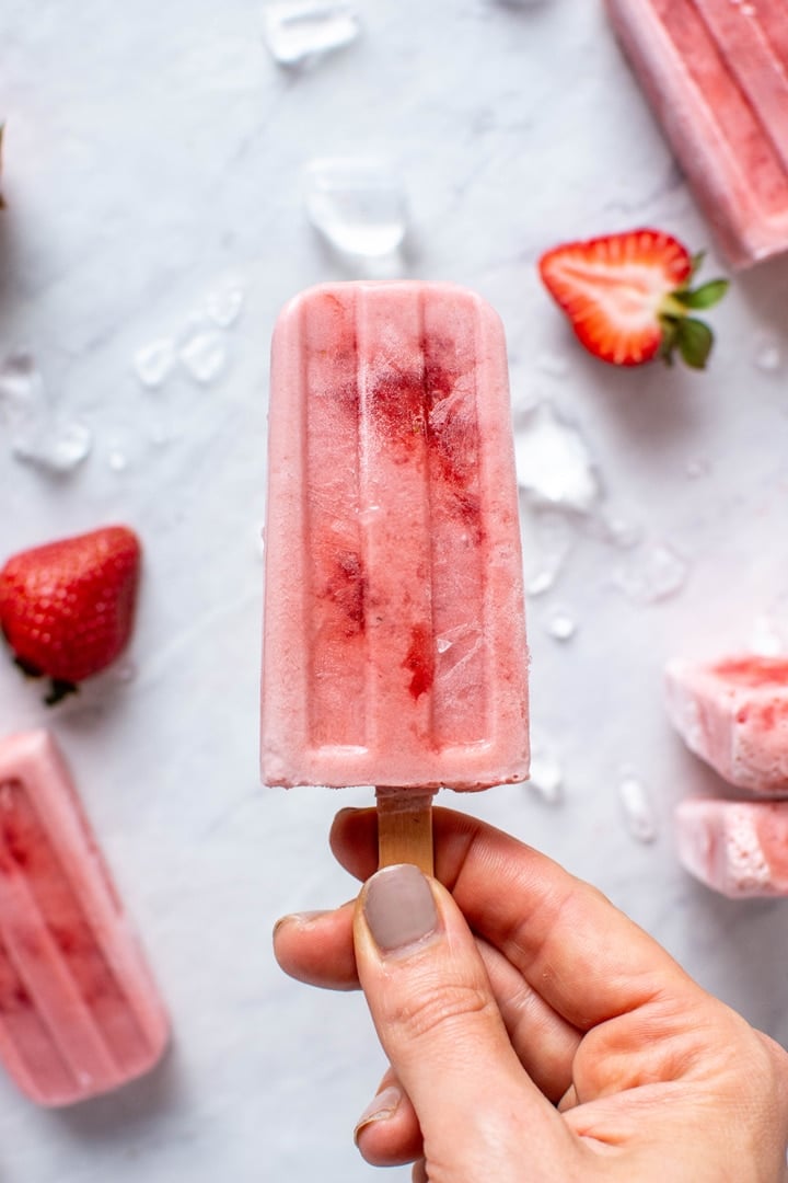 Holding a strawberry coconut milk popsicle against a white background with strawberries
