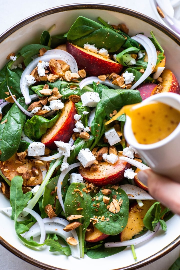 Close up view of a hand pouring a mustardy salad dressing over green salad with peaches, chopped almonds, sliced onion, and crumbled feta cheese