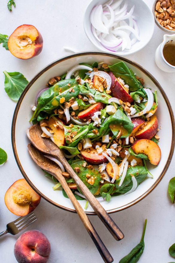 A large bowl filled with green salad with peaches, chopped almonds, sliced onion, and crumbled feta cheese with mustard dressing. Surrounded by peach halves and more salad greens.