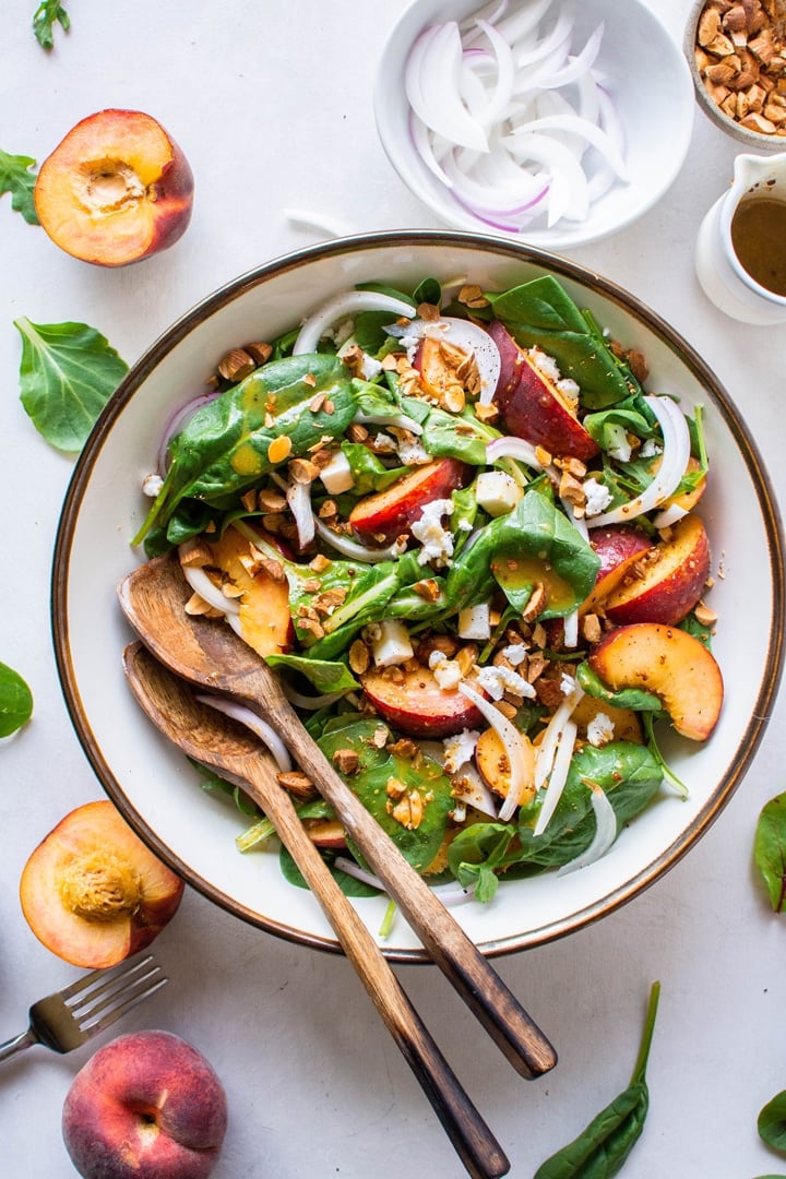 A large bowl filled with green salad with peaches, chopped almonds, sliced onion, and crumbled feta cheese with mustard dressing. Surrounded by peach halves and more salad greens. 