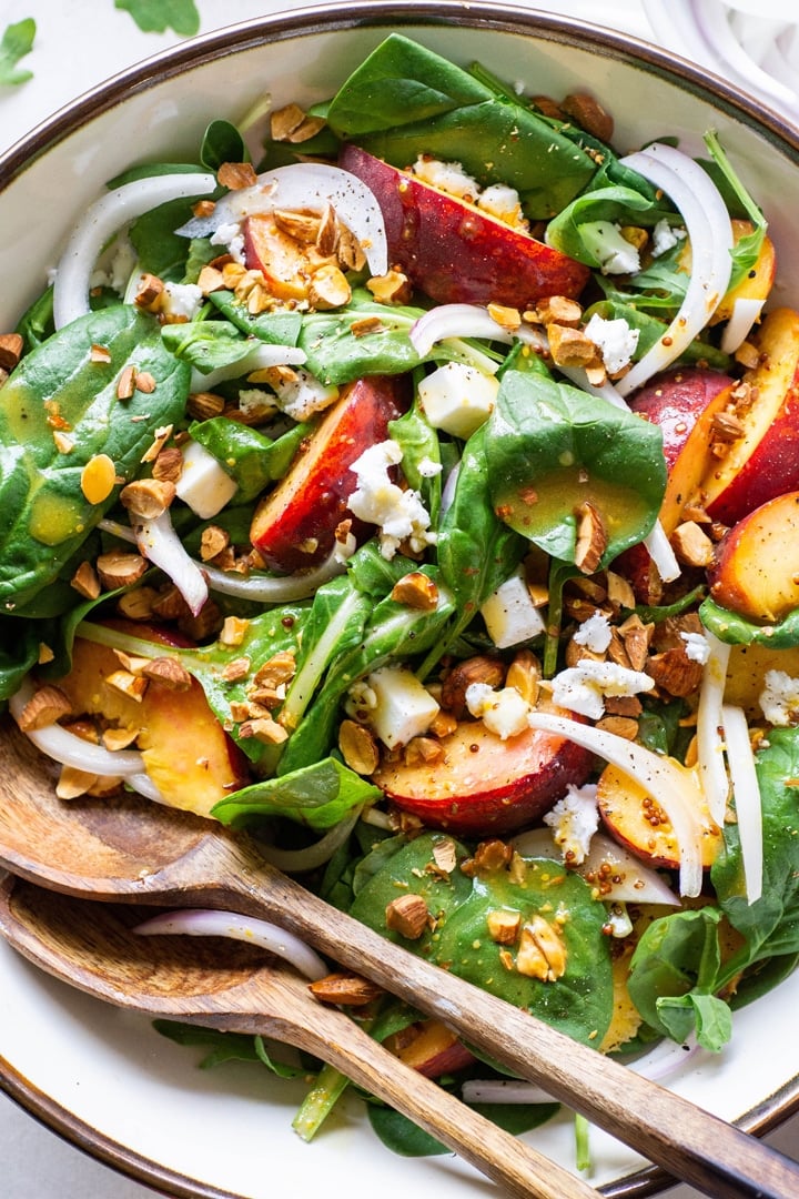 Close up view of a green salad with peaches, chopped almonds, sliced onion, and crumbled feta cheese with mustard dressing