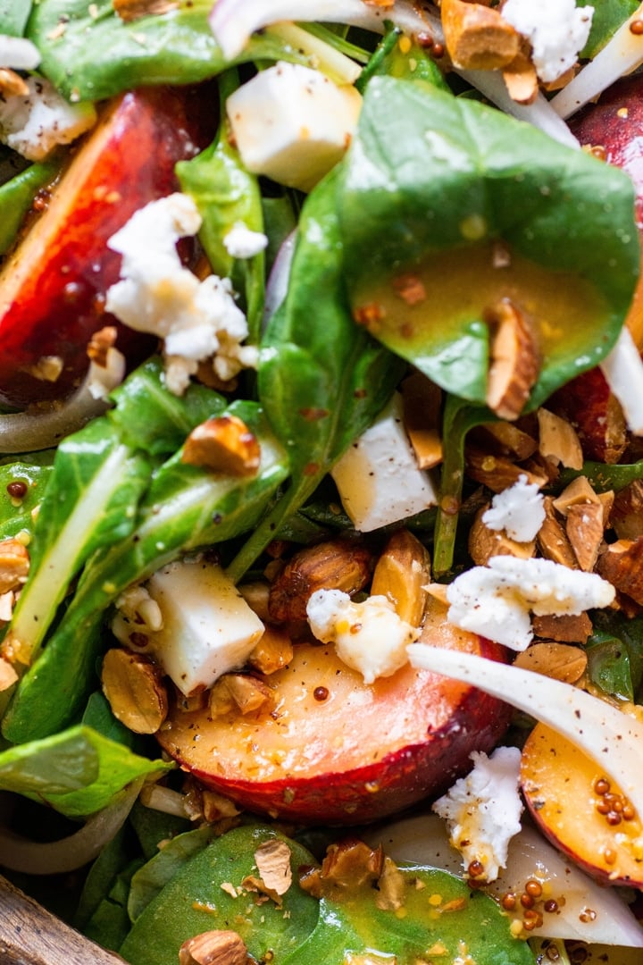 Super close up view of a green salad with peaches, chopped almonds, sliced onion, and crumbled feta cheese with mustard dressing