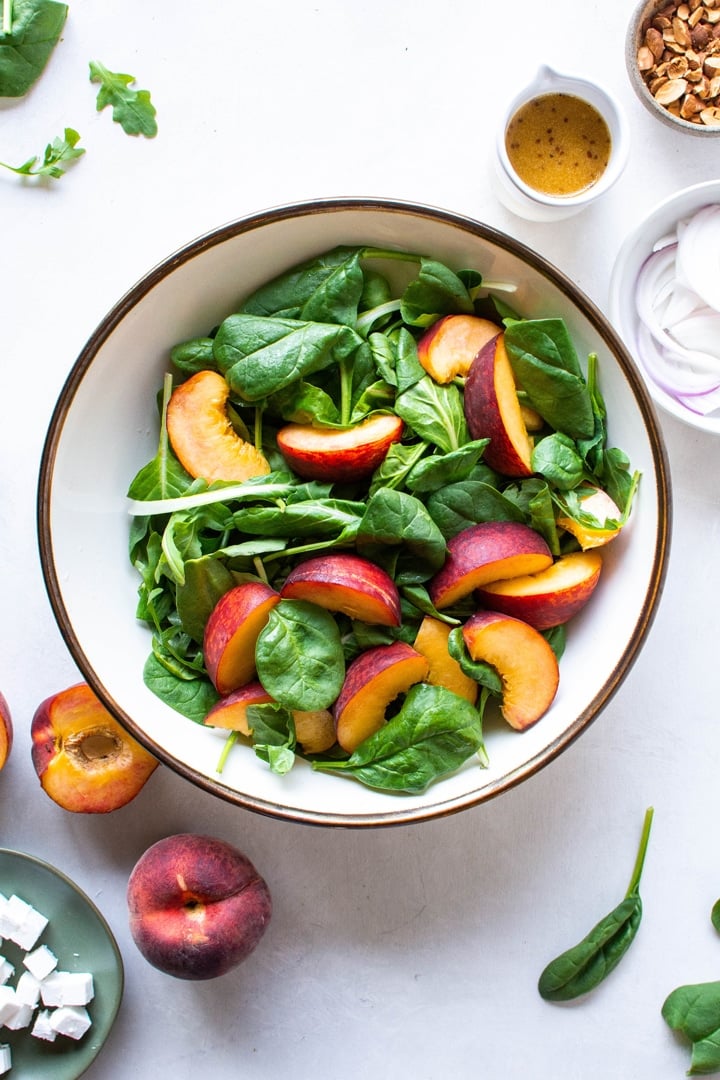 A large bowl of salad greens and sliced peaches on a light colored background surrounded by halved peaches, and a small bowl of red onions. 