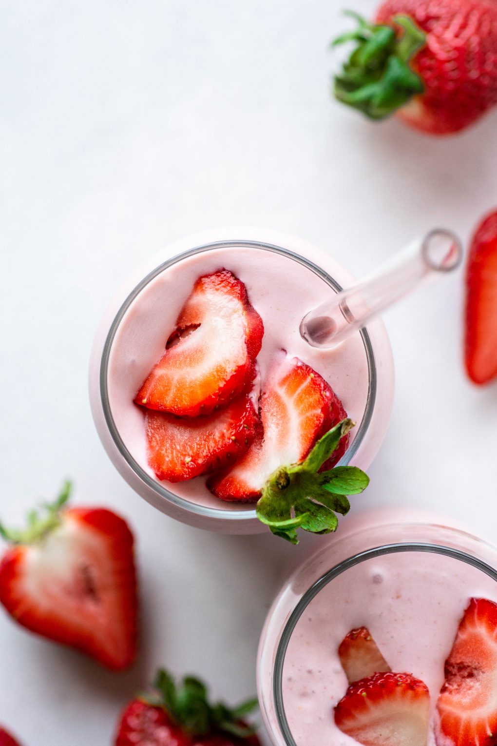 Over head shot of two side by side pink strawberry coconut milk shakes topped with fresh sliced berries on a white background surrounded by more fresh strawberries