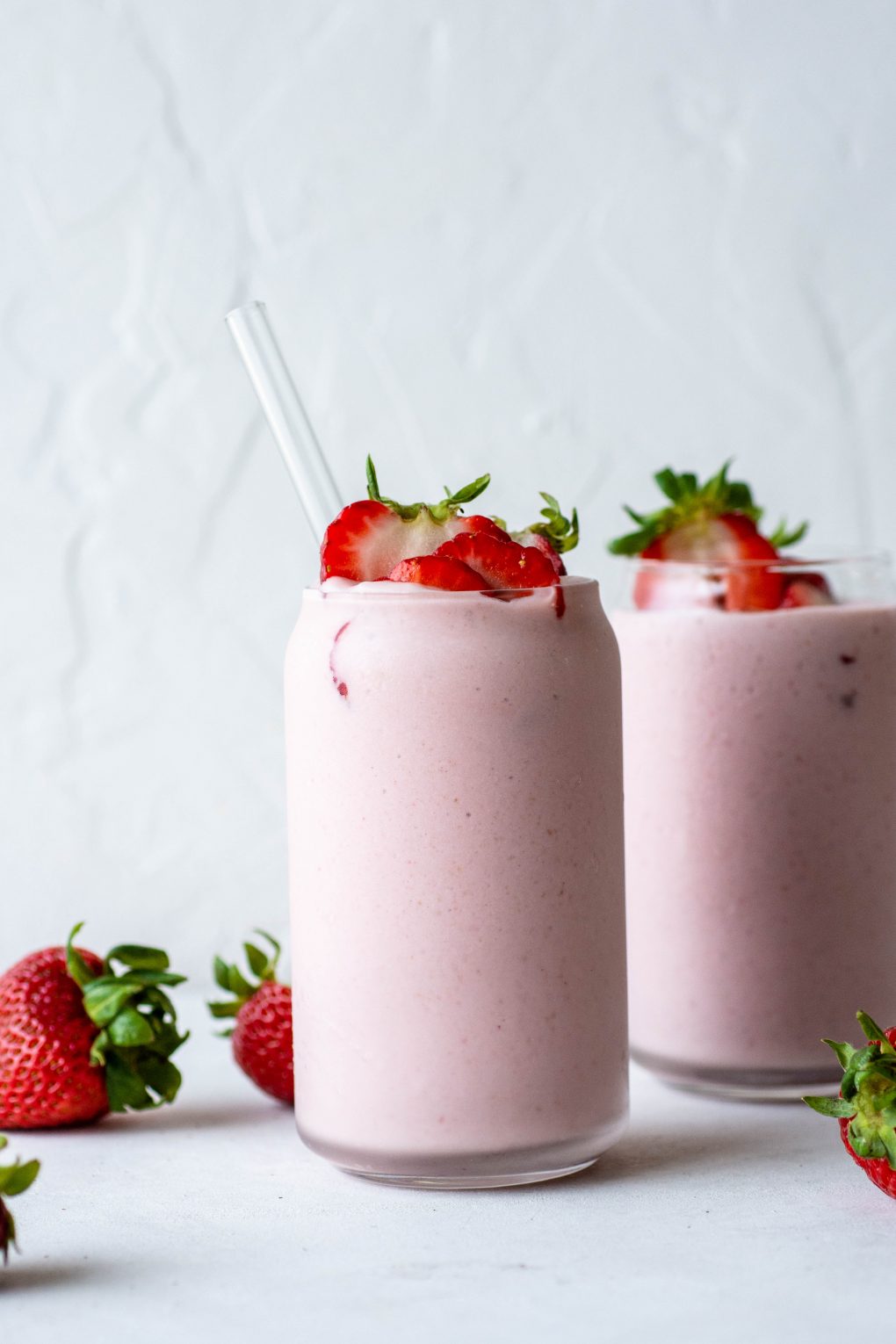 Two side by side thick pink strawberry milkshake from a clear glass jar into a cup topped with fresh strawberries and surrounded by fresh strawberries on a white background