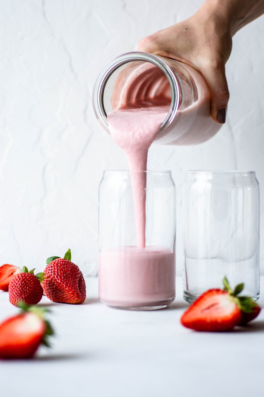 Pouring a thick pink strawberry milkshake from a clear glass jar into a cup surrounded by fresh strawberries on a white background