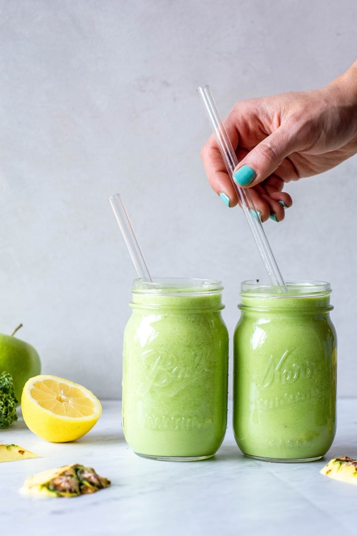 Straight on view of two tropical green smoothies in ball jar glasses with a hand in the right of the frame placing a glass straw in the glass. On a light grey background next to a cut lemon and green apple