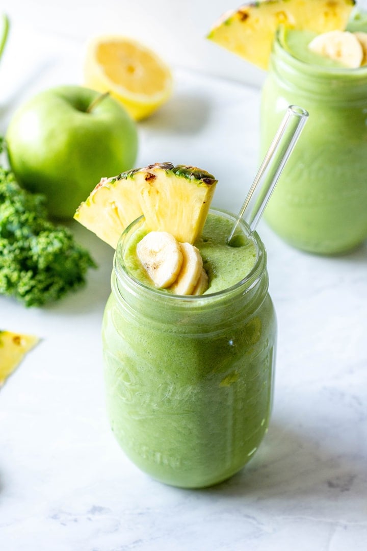 45 degree angle view of a tropical green smoothie topped with some banana slices and a wedge of pineapple. Surrounded by some kale leaves, a green apple, and pineapple peel pieces.