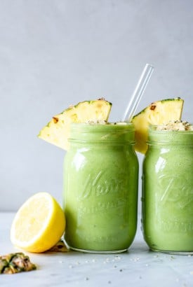 Straight on view of two tropical green smoothies in ball jar glasses with a wedge of pineapple on the rim next to a cut lemon