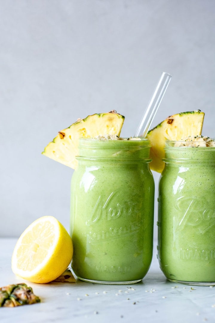 Straight on view of two tropical green smoothies in ball jar glasses with a wedge of pineapple on the rim next to a cut lemon