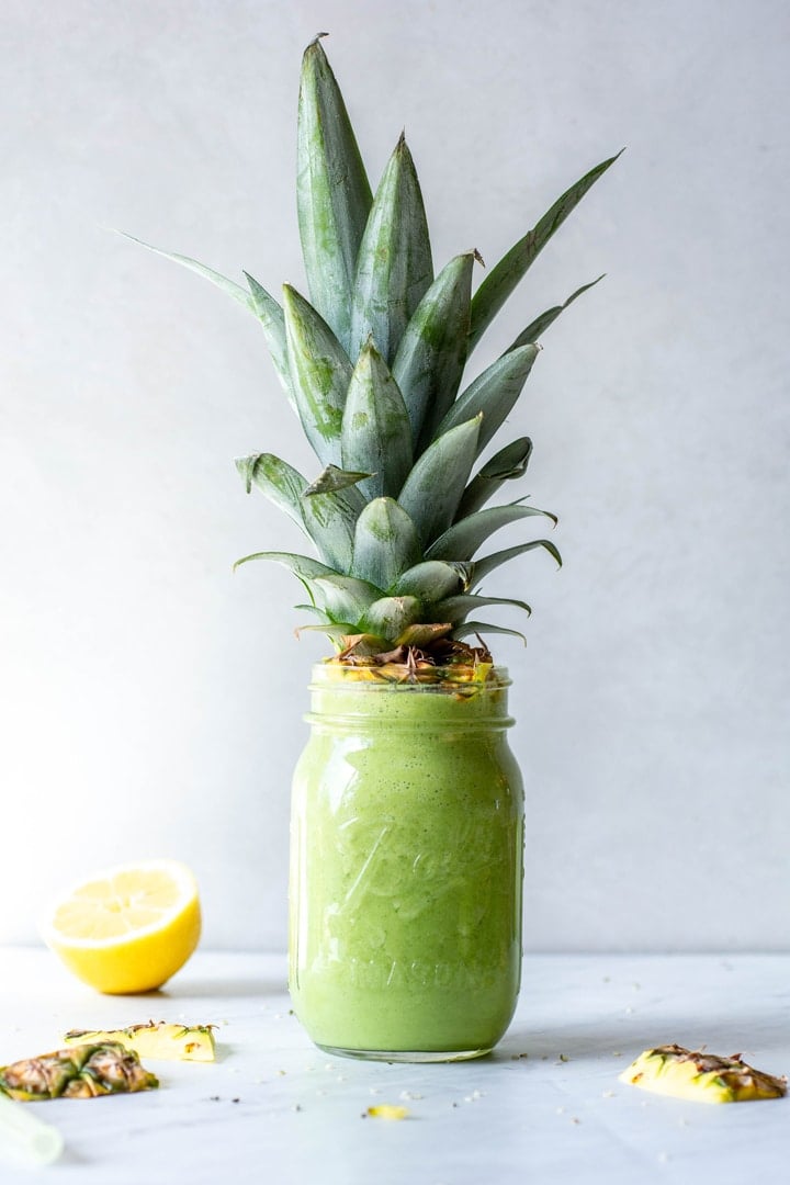 Image of a tropical green smoothie in a ball jar with a pineapple top on the top of the glass. On a light grey background next to a cut lemon and some cut pieces of pineapple peel.