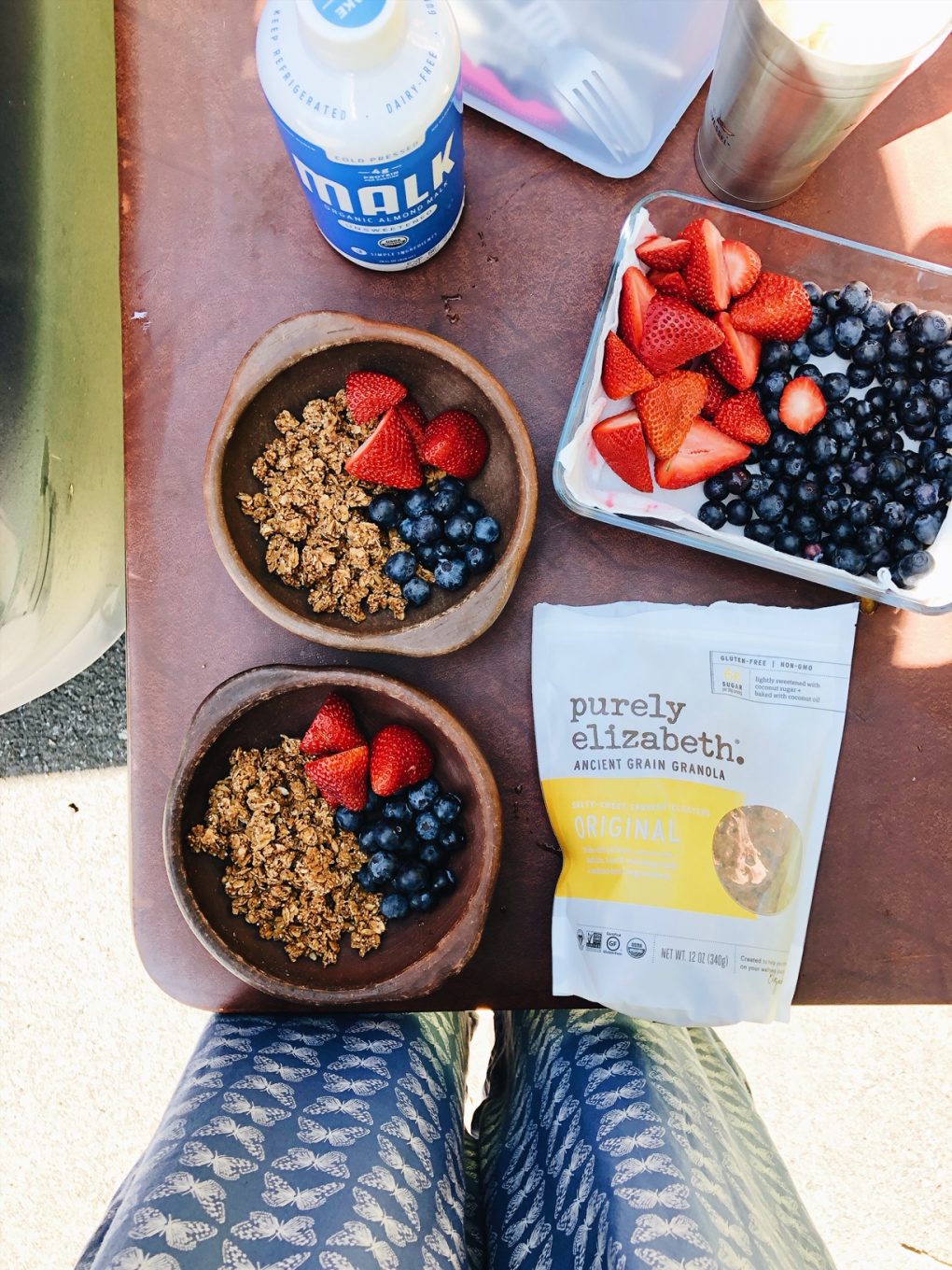 Granola and berries in two small brown bowls  on a brown folding table next to a bowl of berries and a bag of granola