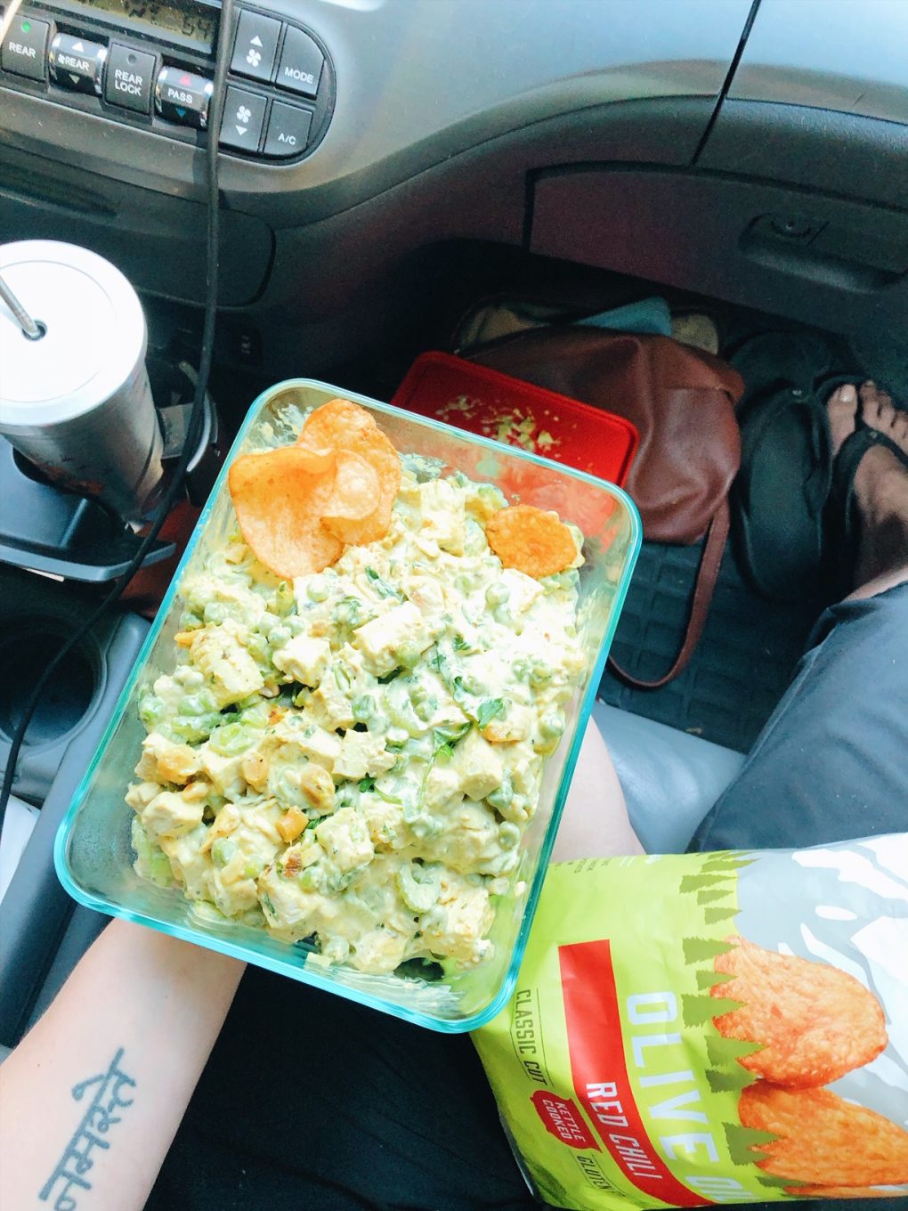 A person sitting in a car holding a glass tupperware with curried chicken salad and potato chips