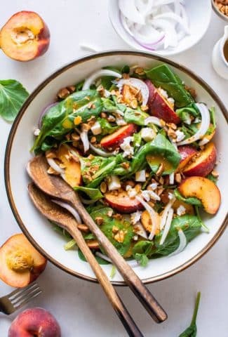 A large bowl filled with green salad with peaches, chopped almonds, sliced onion, and crumbled feta cheese with mustard dressing. Surrounded by peach halves and more salad greens.