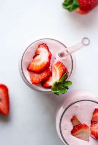 Over head shot of two side by side pink strawberry coconut milk shakes topped with fresh sliced berries on a white background surrounded by more fresh strawberries