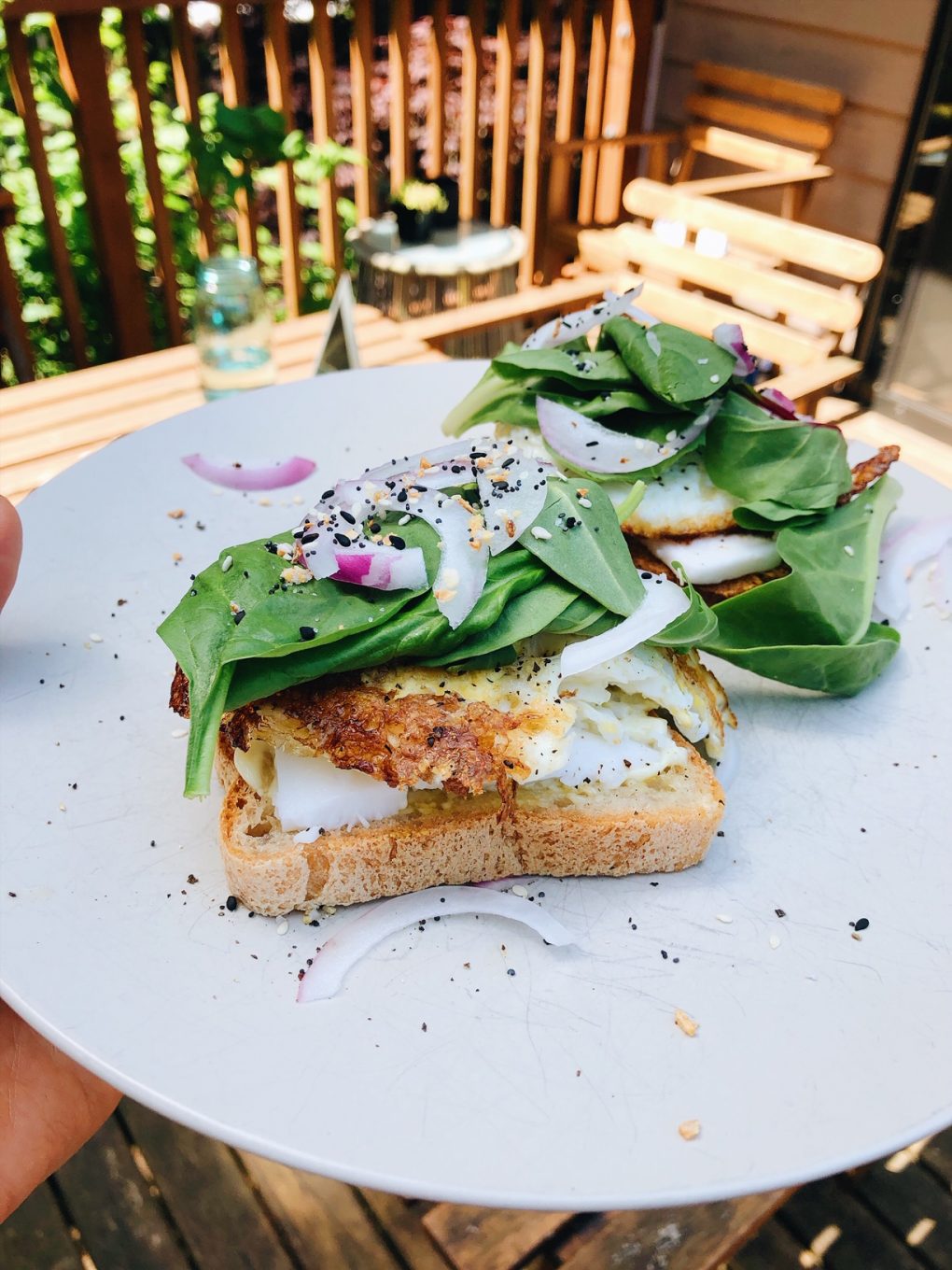 Side view of a plate on an outdoor wooden table with two open faced fried egg sandwiches with baby spinach, red onion, and everything bagel spice