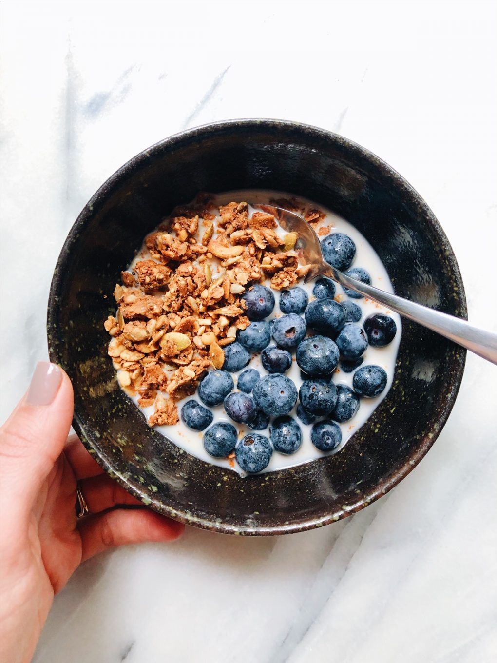 Granola and blueberries in a small black bowl held by a hand in the left of the frame on a white background