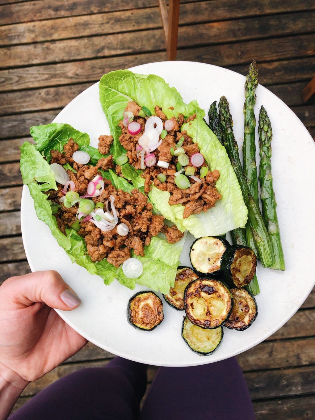 Holding a plate with chicken lettuce wraps with a side of asparagus and zuchinni outside on a wooden deck. 
