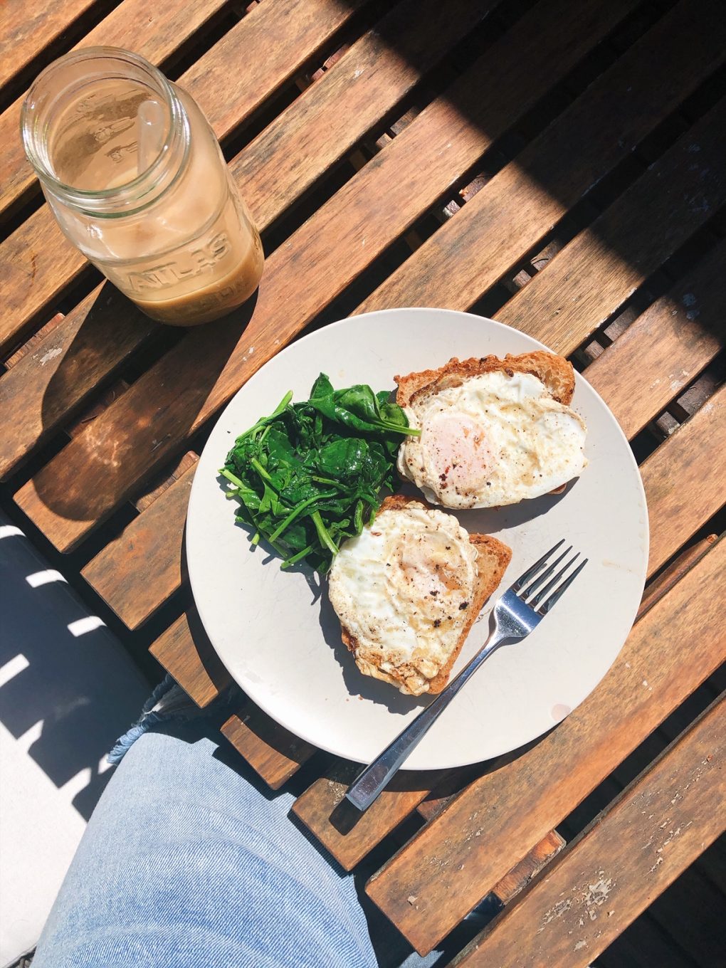 A plate on an outdoor wooden table with two open faced fried egg sandwiches and a side of sauteed spinach