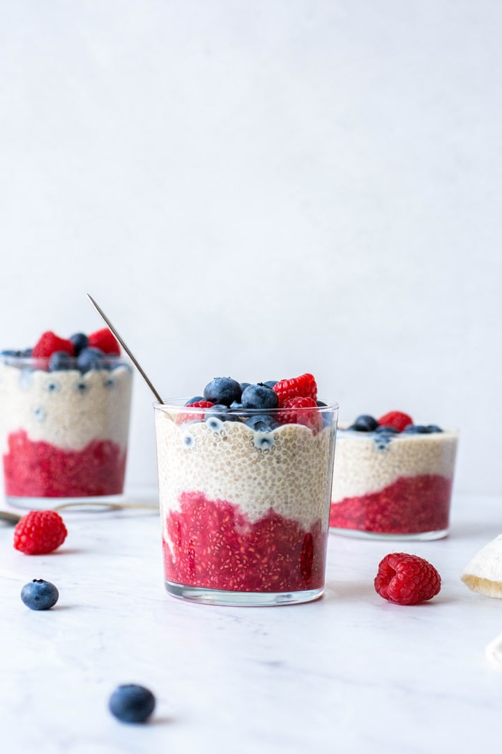 Side view of 3 staggered glass cups with layers of chia seed pudding - raspberry, vanilla, and fresh blueberries on top. Surrounded by fresh berries on a white background