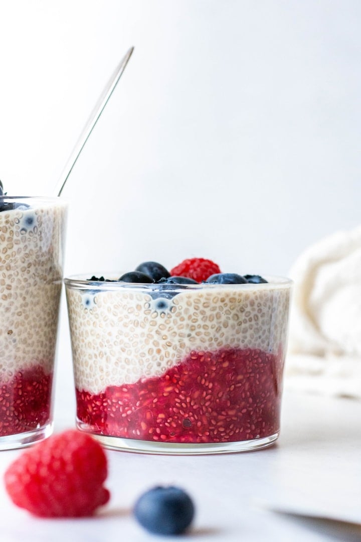 Close up side view of a small glass cup with layered chia seed pudding topped with blueberries and raspberries on a white background