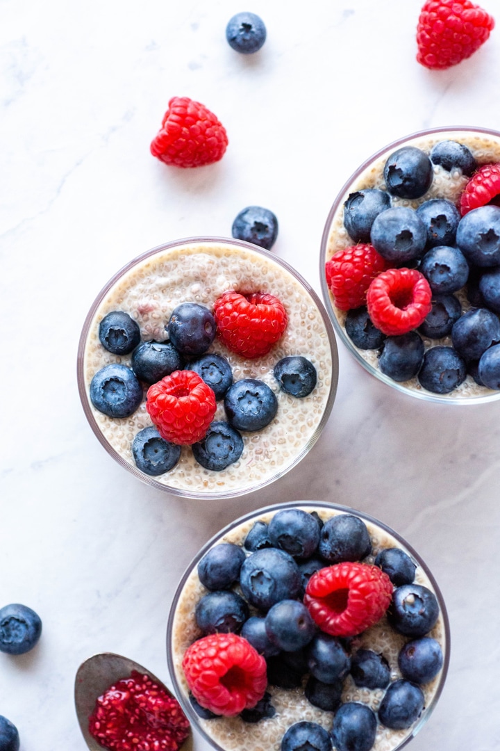 Overhead shot of three round cups of chia vanilla chia seed pudding topped with fresh raspberries and blueberries. On a white background surrounded by a few scattered berries. 