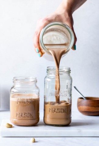 Pouring chocolate cashing milk into a tall mason jar next to an already filled mason jar on a white background