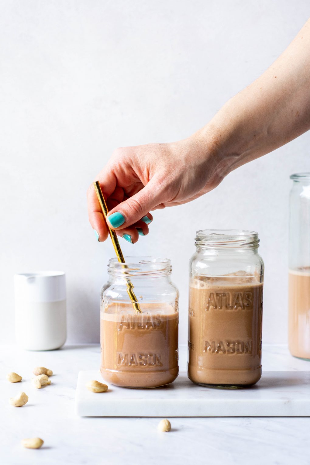 Placing a straw in one of two side by side glasses of chocolate cashew milk on a white background