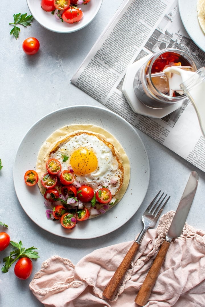 Two plates with open faced fried egg and hummus breakfast tacos topped with a simple tomato salad. Sitting on top of a newspaper on a light colored background next to a fork and knife, and someone pouring milk into an iced coffee. 