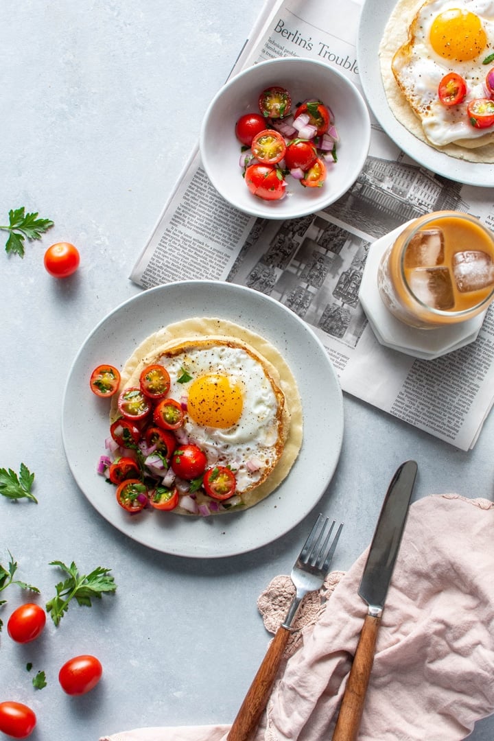Overhead view of a plate with open faced fried egg and hummus breakfast taco topped with a simple tomato salad. Sitting on top of a newspaper on a light colored background next to an iced coffee, a fork and knife, and a small bowl with extra tomato salad.