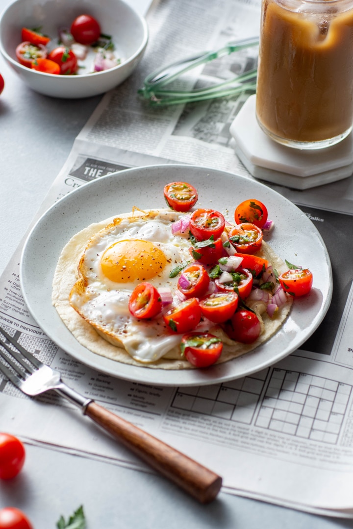 Side angle of a plate with open faced fried egg and hummus breakfast taco topped with a simple tomato salad. Sitting on top of a newspaper on a light colored background next to an iced coffee and some cute sunglasses.