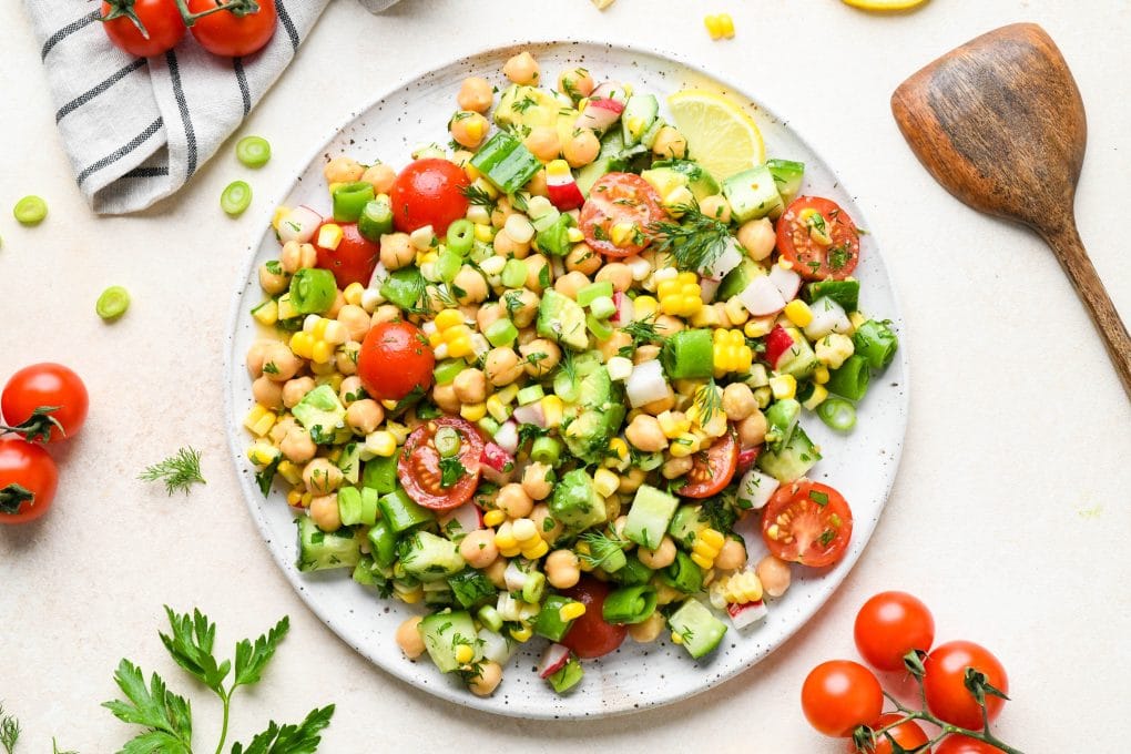 Vegan chickpea salad on a large round white speckled plate surrounded by fresh ingredients and a striped linen.