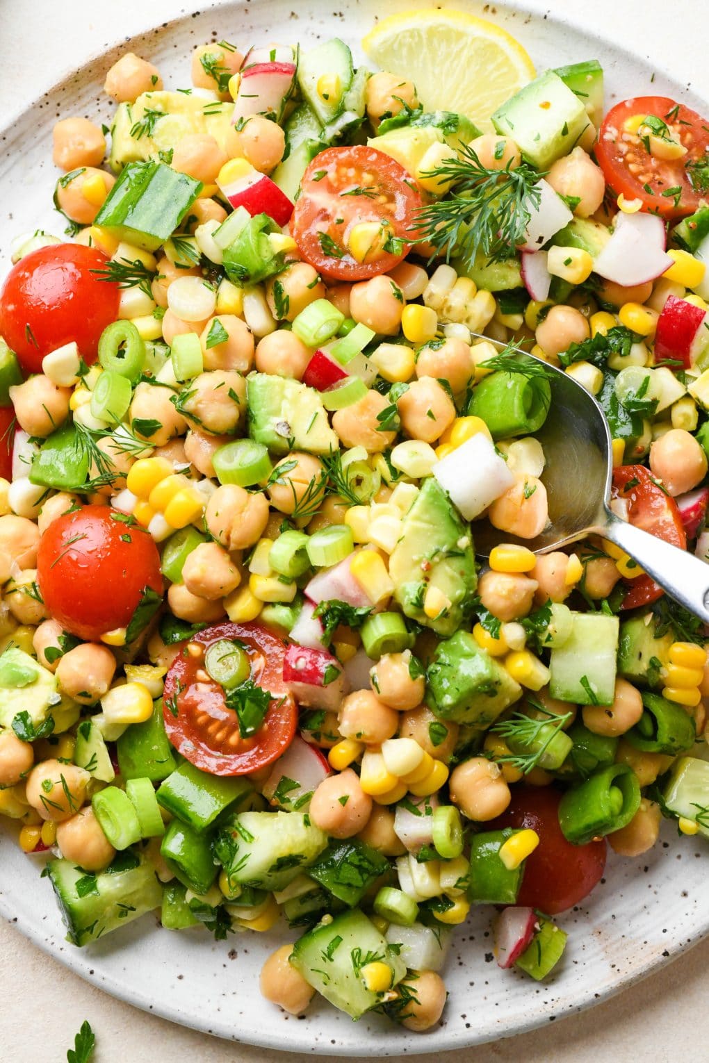 Colorful vegan chickpea salad on a large white speckled plate. Topped with fresh herbs and lemon wedges.