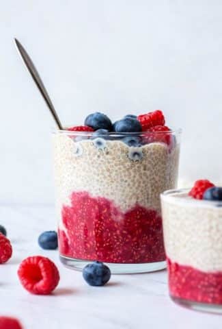 Side view of a glass cup with layers of chia seed pudding - raspberry, vanilla, and fresh blueberries on top. Surrounded by fresh berries on a white background