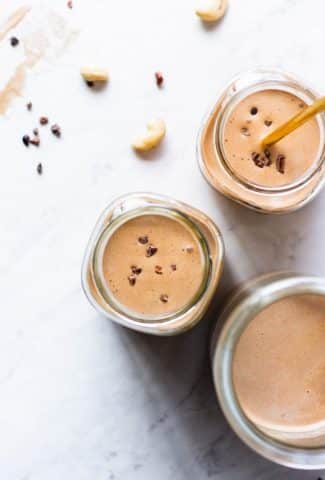 Over head shot of three glasses of chocolate cashew milk on a white background surrounded by a few cashews and some cacao nibs