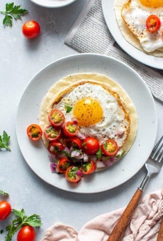 Two plates with open faced fried egg and hummus breakfast tacos topped with a simple tomato salad. Sitting on top of a newspaper on a light colored background next to a fork and knife.