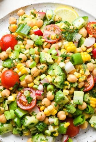 Colorful vegan chickpea salad on a large white speckled plate. Topped with fresh herbs and lemon wedges.