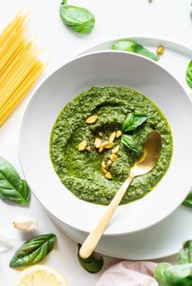 Overhead view of a white bowl of pistachio basil pesto topped with fresh basil and pistachios on a white background next to scattered basil leaves and a bundle of uncooked spaghetti