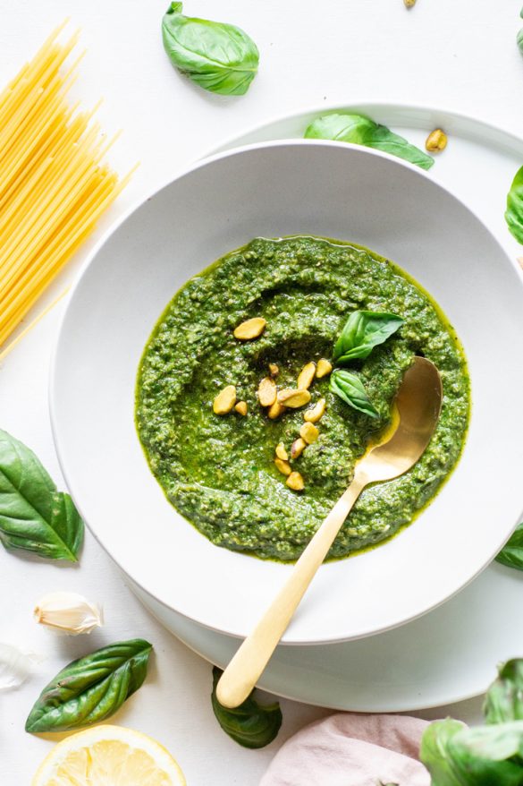 Overhead view of a white bowl of pistachio basil pesto topped with fresh basil and pistachios on a white background next to scattered basil leaves and a bundle of uncooked spaghetti