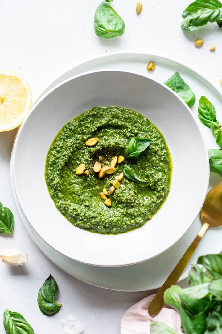 Overhead view of a white bowl of pistachio basil pesto topped with fresh basil and pistachios on a white background next to scattered basil leaves and a lemon