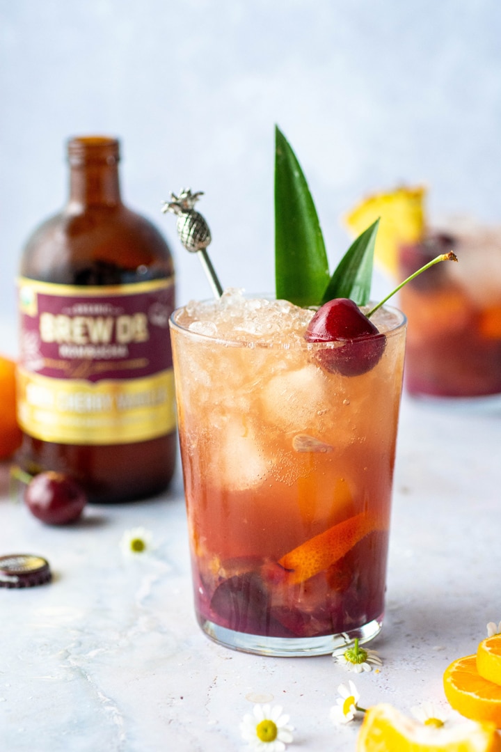 A tropical cocktail with pineapple leaves and a cherry on a light blue background in front of a bottle of Brew Dr. Dark cherry vanilla Kombucha