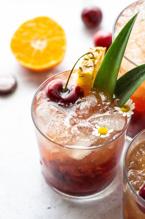 Side view of three tropical kombucha cocktails next to each other with pineapple leaves, pineapple wedges, dark cherries, and sliced tangerine on a light background