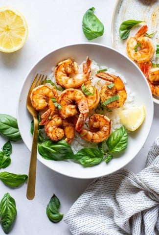 Large white bowl with white rice topped with seared shrimp with fresh basil and lemon wedges on a light background