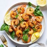 Plate of seared shrimp topped with fresh basil and lemon wedges on a light background next to loose basil leaves, a fork, and a striped dish towel