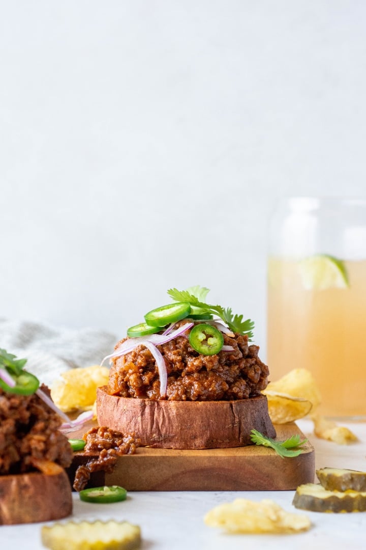 Side angle shot of two open faced sweet potato sloppy joe sandwiches topped with cilantro, red onion, and jalapeno against a white background
