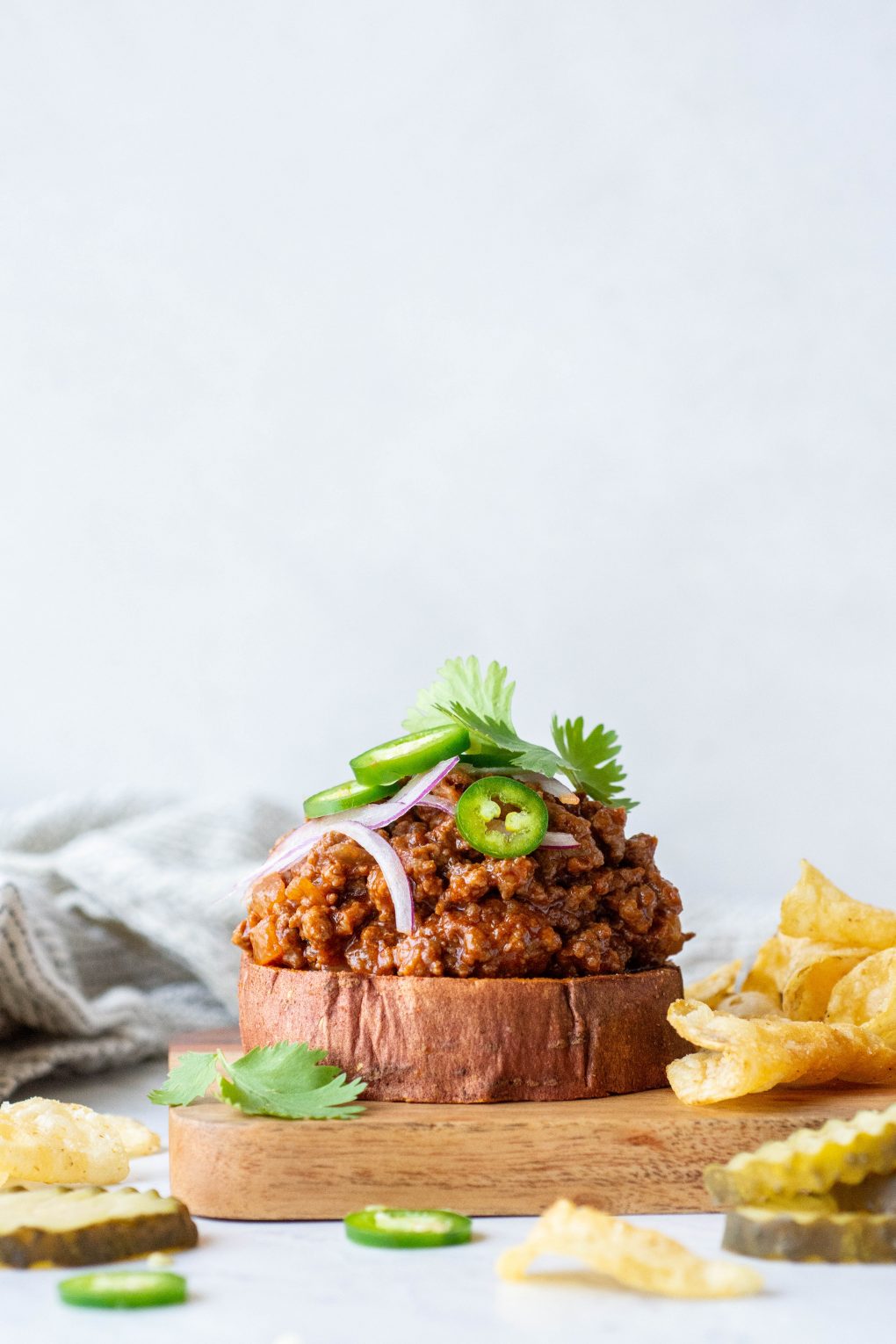 Side angle shot of a open faced sweet potato sloppy joe sandwich topped with cilantro, red onion, and jalapeno against a white background on a wooden board next to potato chips and pickle slices
