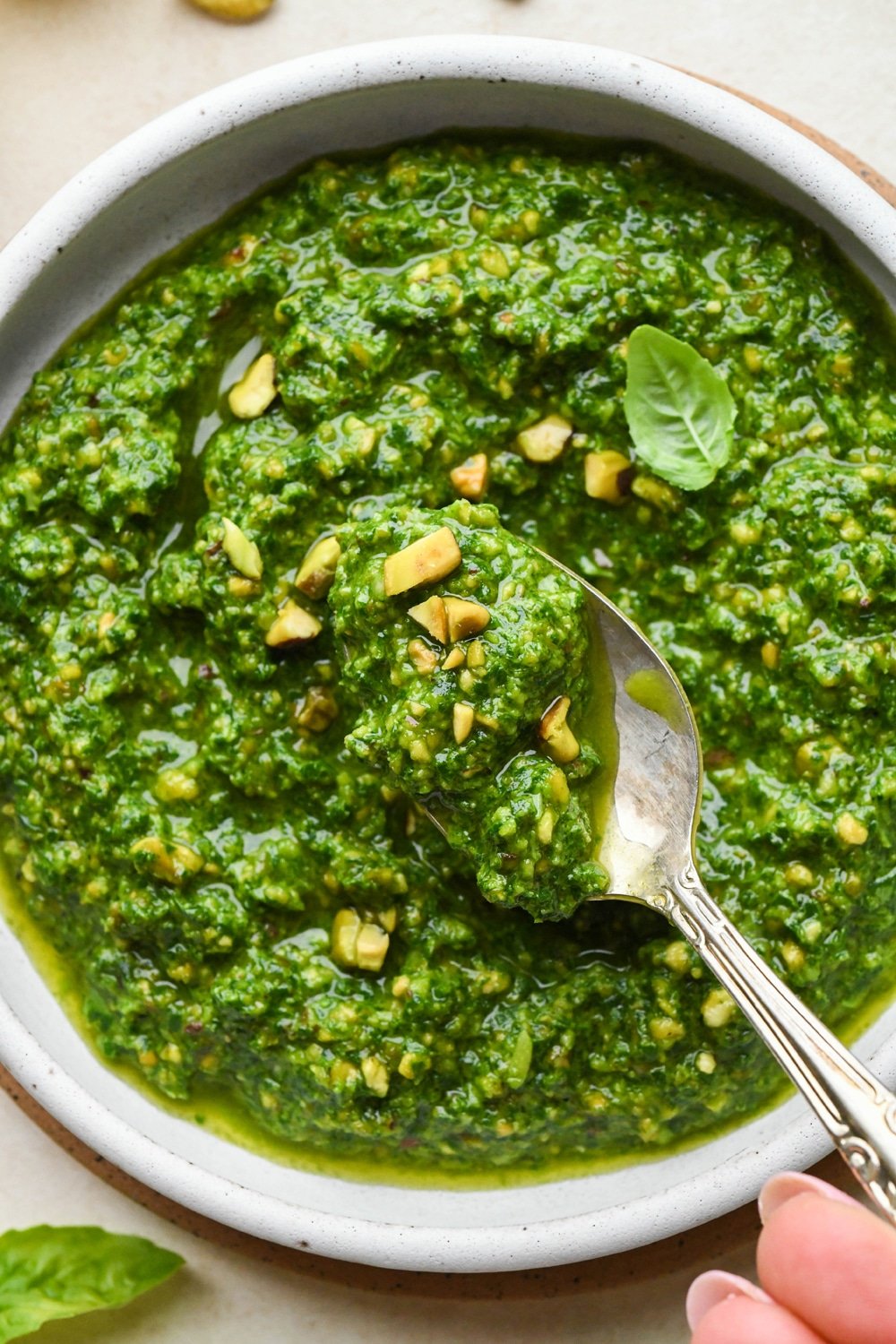 A small bowl of bright green pistachio pesto, topped with a few chopped pistachios and a small basil leaf, with a teaspoon lifting our a small portion of pesto.