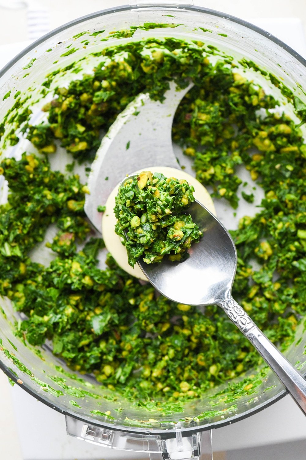 How to make pistachio pesto: Dry ingredients in the container of a food processor after pulsing together, with a spoon lifting some of the mixture out to show the texture.