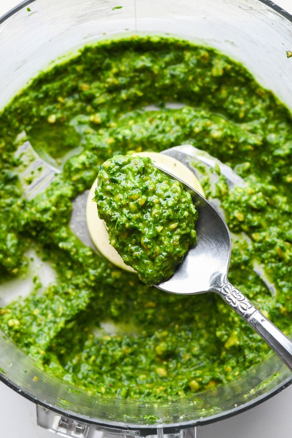 How to make pistachio pesto: A spoon lifting out a spoonful of creamy pesto in food processor container after adding olive oil.