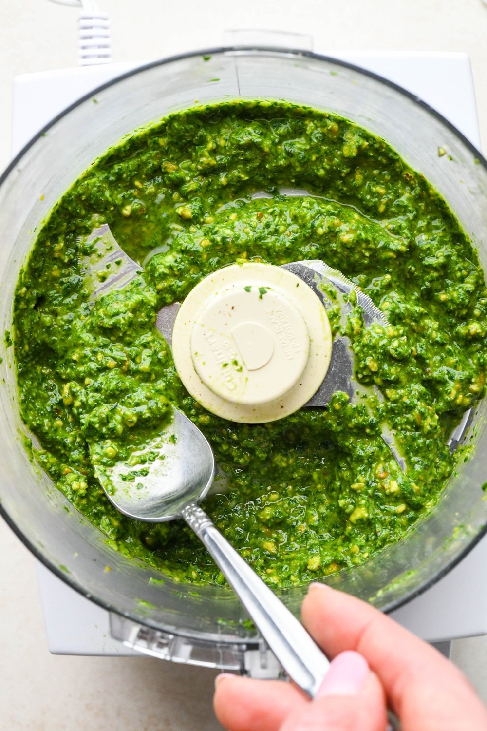 How to make pistachio pesto: A spoon angled into food processor container with creamy pesto after adding olive oil.